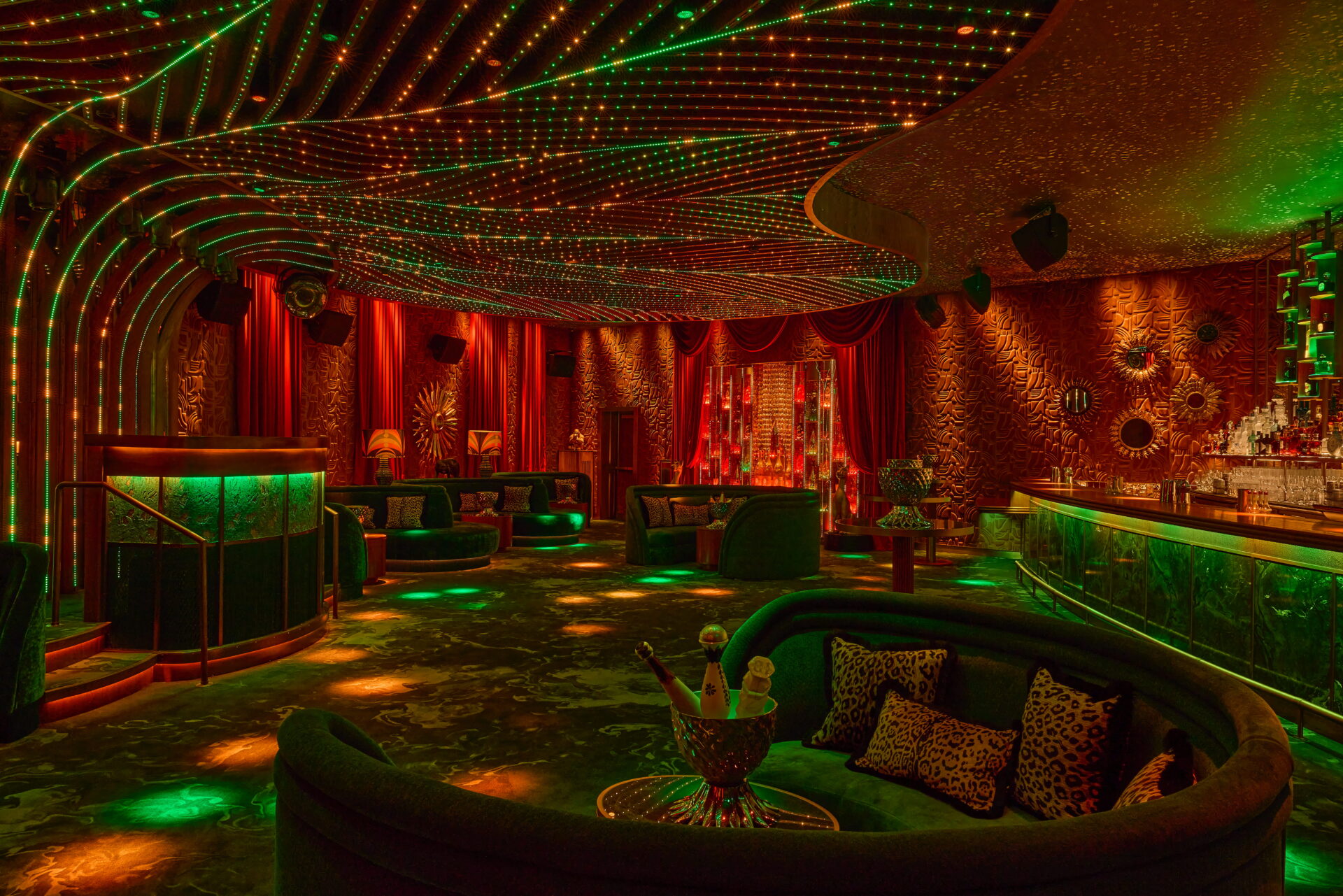 A panoramic view of the Selva nightclub interior at Amazónico, featuring curved green velvet booths with leopard-print cushions. The ceiling displays a canopy of undulating green and amber fibre-optic lights. Textured red walls are decorated with golden sunburst mirrors, while a glowing green bar and DJ podium frame the room. The space is finished with dark patterned flooring and warm red accent lighting.