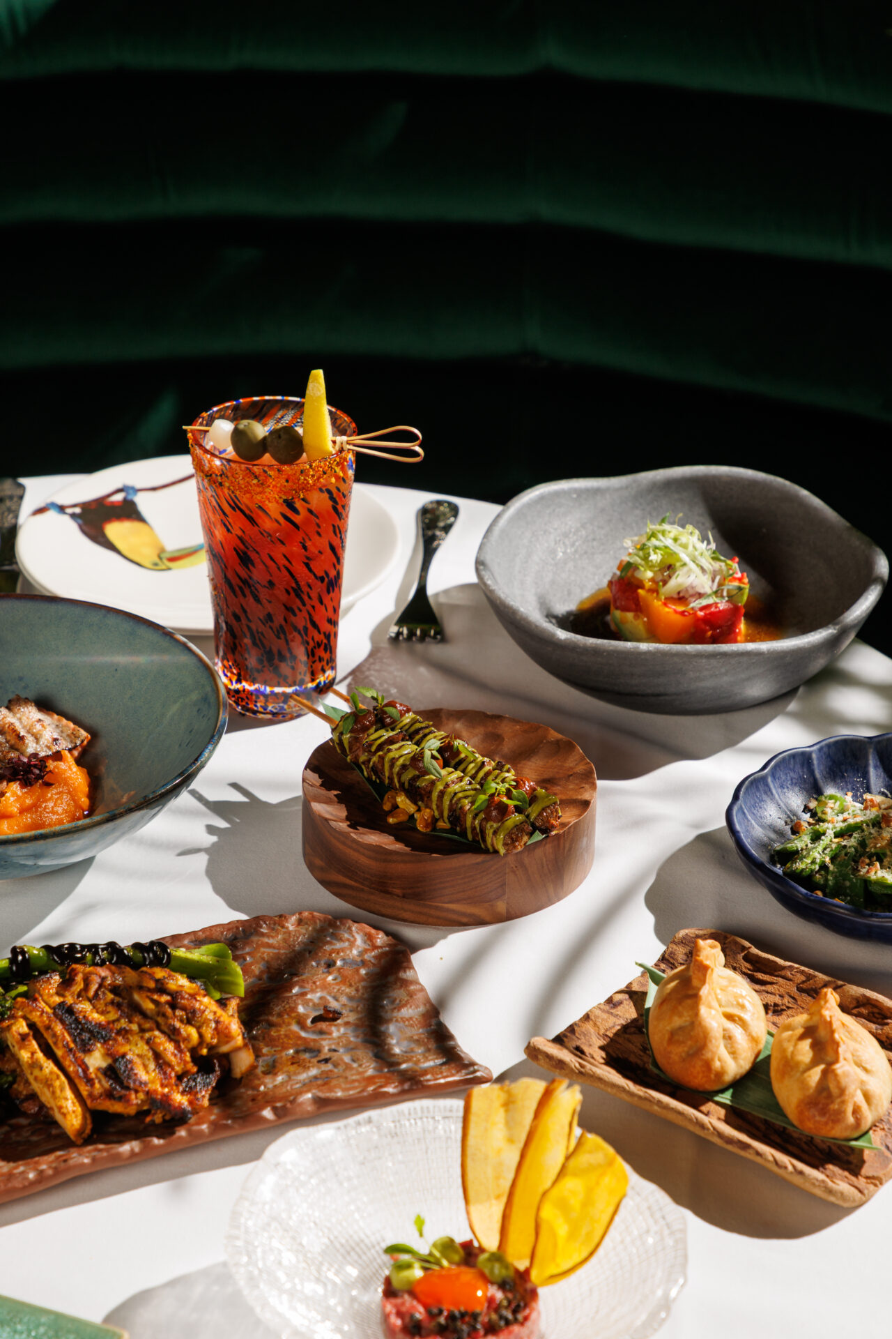 A dramatic sunlit spread of Amazónico set lunch dishes including a Bloody Mary, meat skewers, dumplings, tartare, and grilled chicken, set against deep green velvet banquette seating.