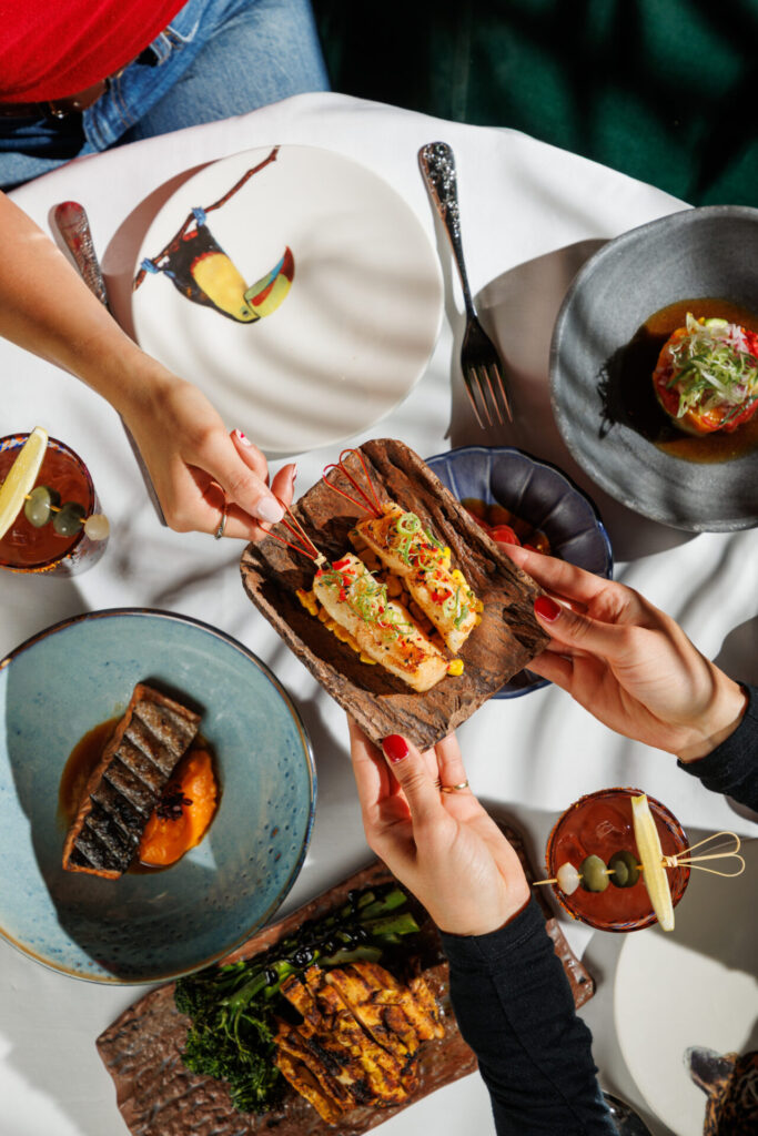 Two guests sharing grilled fish skewers across a sunlit Amazónico lunch table, with Bloody Marys, grilled fish, and sharing plates on illustrated toucan crockery.