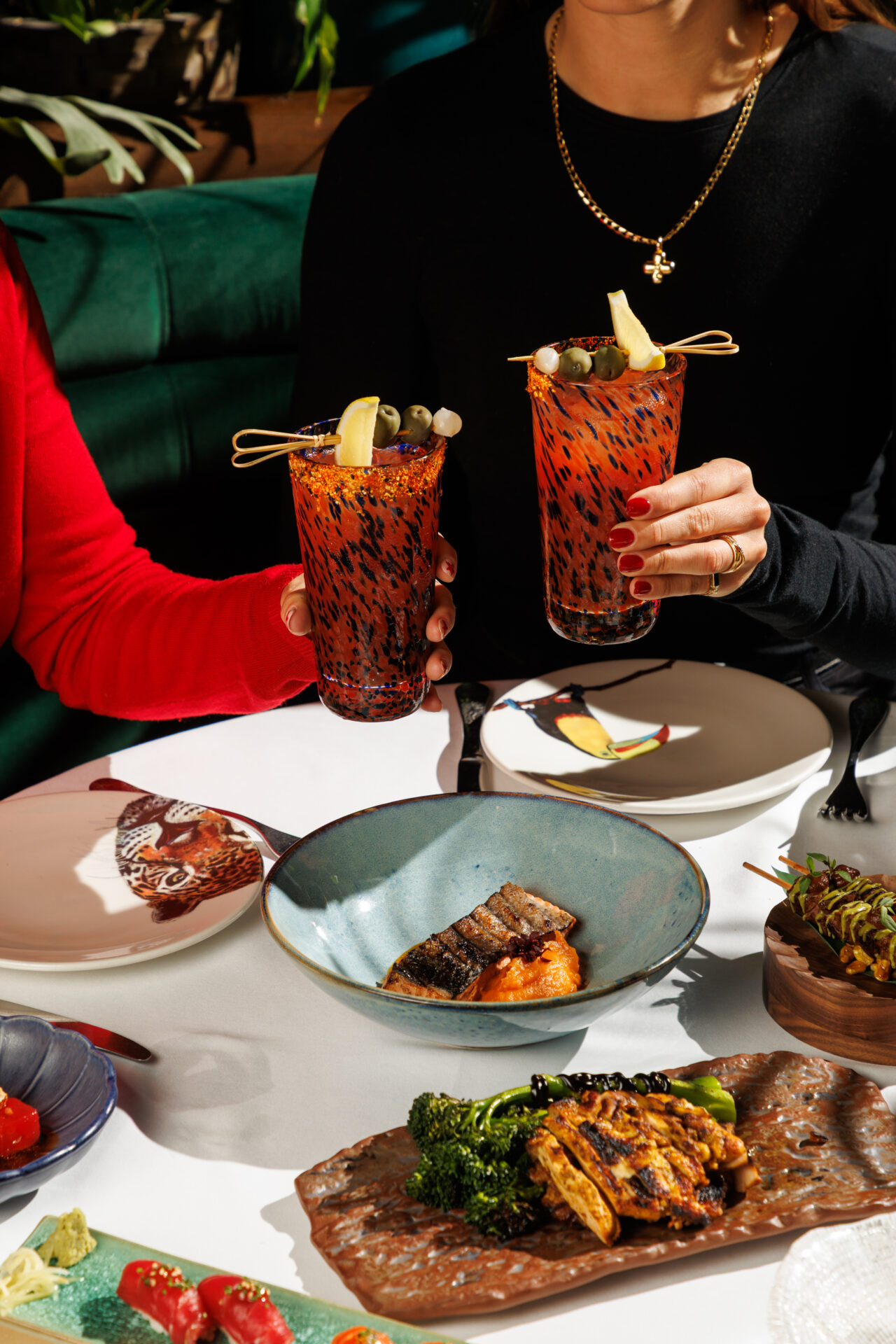 Two guests toasting with Bloody Mary cocktails at a vibrant Amazónico lunch table, surrounded by grilled fish, sushi, and sharing plates in the sun-filled restaurant.