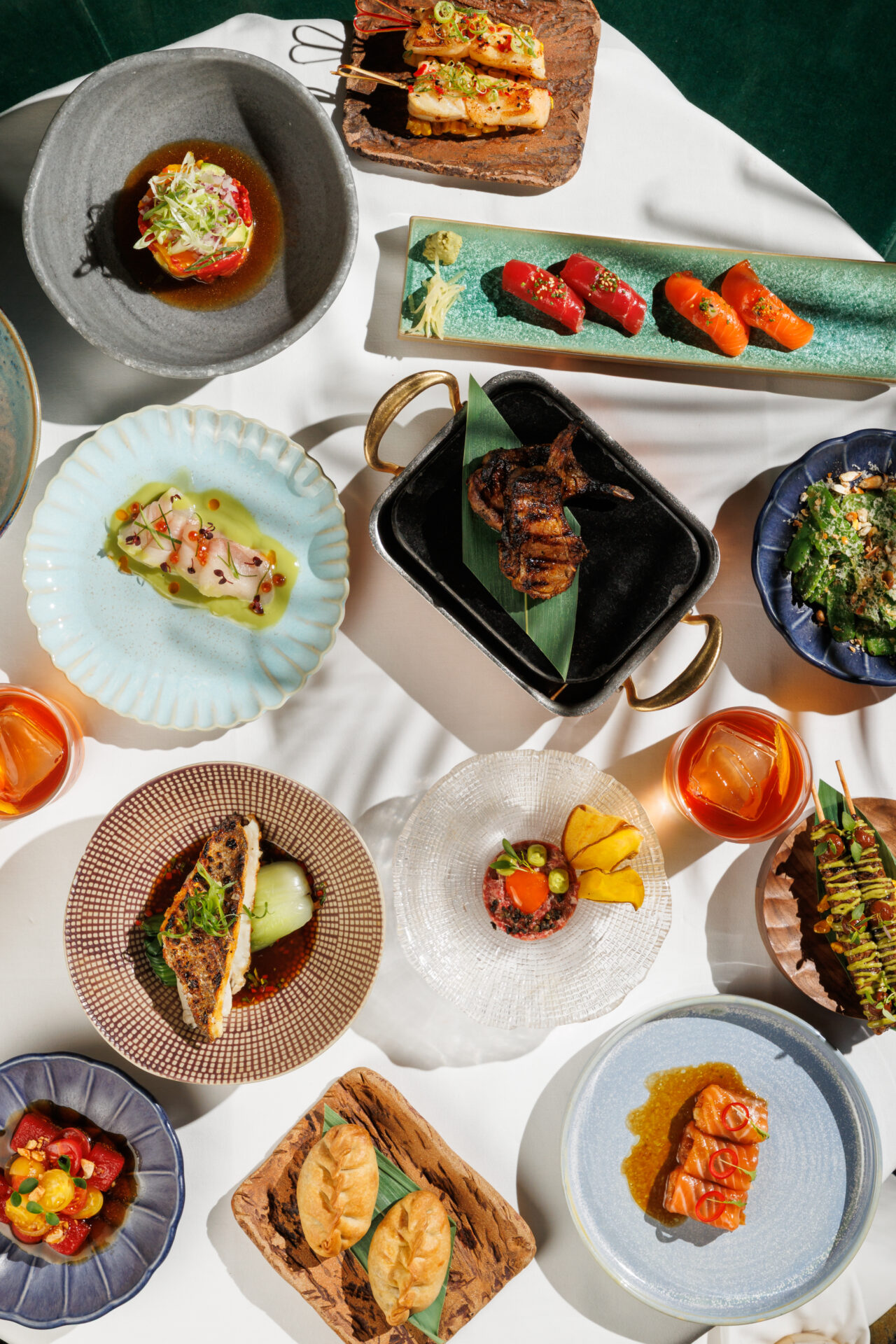 An abundant overhead spread of Amazónico's set lunch dishes, including sushi, grilled fish, tartare, bao buns, and cocktails, served on eclectic artisan plates and boards.