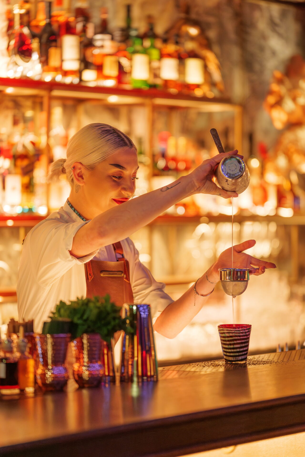 Selva bartender crafting a cocktail behind the bar, surrounded by colourful glassware and illuminated spirits in the nightclub’s mysterious, wild setting.