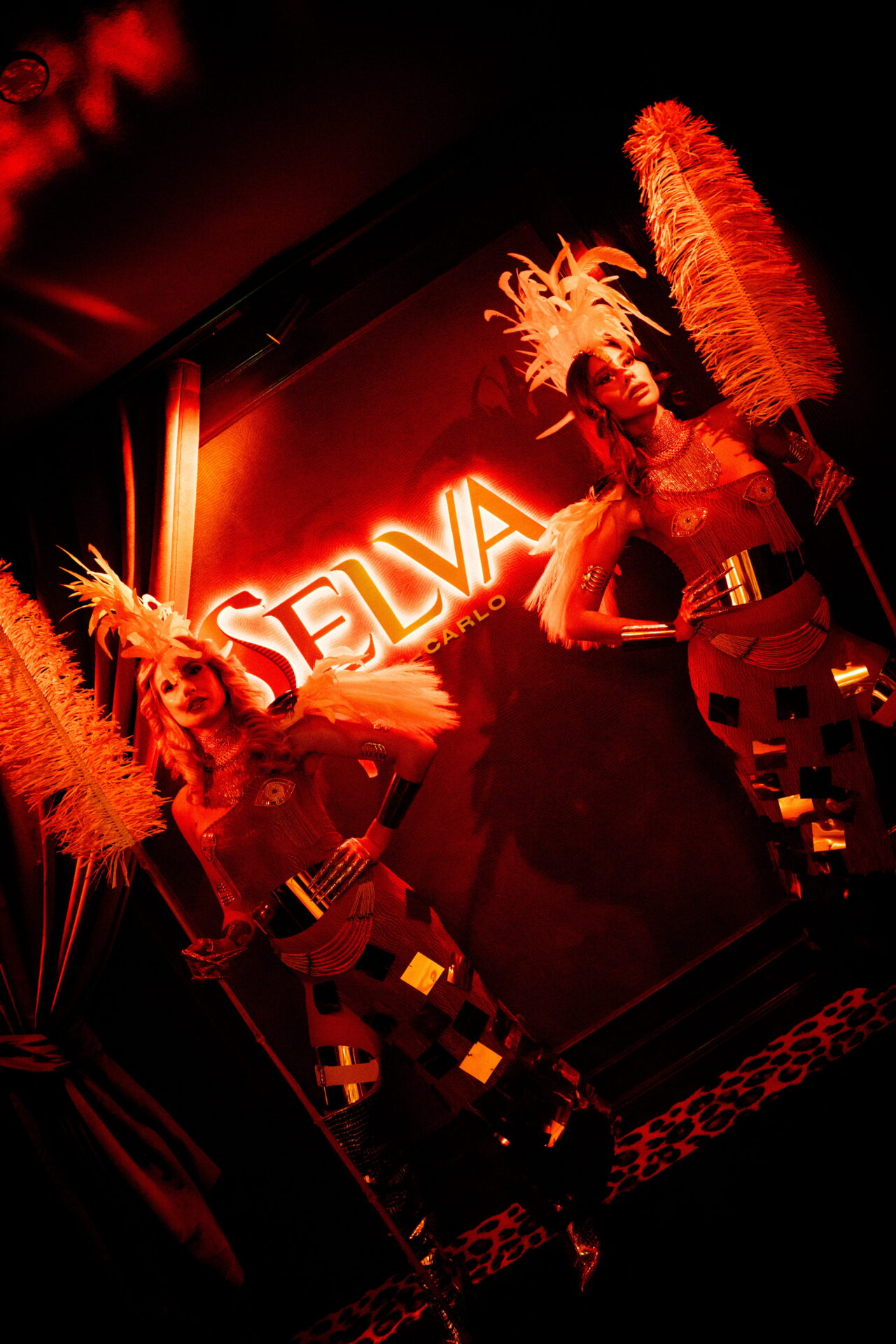 Selva Monte Carlo entrance coming alive after dark, with a neon Selva sign, glowing red and amber lights, and two dancers with feather costumes and an intimate atmosphere.